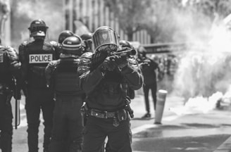 A group of police officers in riot gear stands in a smoke-filled urban environment. The central officer holds a baton or similar object, with other officers visible in the background. Their uniforms are protective and tactical, indicative of a high-tension situation. The scene is monochromatic, emphasizing the seriousness and dramatic nature of the event.
