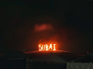 The word 'DUBAI' is illuminated in bright, fiery lights on a hillside, against a dark night sky. The scene has a dramatic and captivating appearance with tents or rooftops visible at the bottom edge of the image.