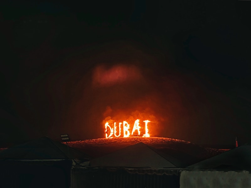 The word 'DUBAI' is illuminated in bright, fiery lights on a hillside, against a dark night sky. The scene has a dramatic and captivating appearance with tents or rooftops visible at the bottom edge of the image.