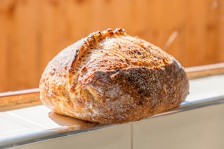 A freshly baked loaf of bread with a rustic crust, resting on a tiled surface. The bread has a golden-brown, crispy exterior with visible flour dusting and charred marks.