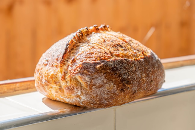 A freshly baked loaf of bread with a rustic crust, resting on a tiled surface. The bread has a golden-brown, crispy exterior with visible flour dusting and charred marks.