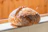 A freshly baked loaf of bread with a rustic crust, resting on a tiled surface. The bread has a golden-brown, crispy exterior with visible flour dusting and charred marks.