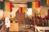 A rustic room filled with colorful yarns hanging from the ceiling, giving a vibrant look. There are various wooden weaving looms and vintage sewing machines scattered throughout the space. The walls appear aged and textured, accentuating the workshop-like atmosphere. There's a small table covered with more yarns, fabric, and plants, adding to the creative ambiance.