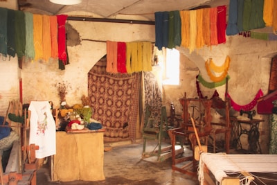 A rustic room filled with colorful yarns hanging from the ceiling, giving a vibrant look. There are various wooden weaving looms and vintage sewing machines scattered throughout the space. The walls appear aged and textured, accentuating the workshop-like atmosphere. There's a small table covered with more yarns, fabric, and plants, adding to the creative ambiance.