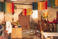 A rustic room filled with colorful yarns hanging from the ceiling, giving a vibrant look. There are various wooden weaving looms and vintage sewing machines scattered throughout the space. The walls appear aged and textured, accentuating the workshop-like atmosphere. There's a small table covered with more yarns, fabric, and plants, adding to the creative ambiance.