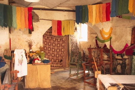 A rustic room filled with colorful yarns hanging from the ceiling, giving a vibrant look. There are various wooden weaving looms and vintage sewing machines scattered throughout the space. The walls appear aged and textured, accentuating the workshop-like atmosphere. There's a small table covered with more yarns, fabric, and plants, adding to the creative ambiance.