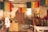 A rustic room filled with colorful yarns hanging from the ceiling, giving a vibrant look. There are various wooden weaving looms and vintage sewing machines scattered throughout the space. The walls appear aged and textured, accentuating the workshop-like atmosphere. There's a small table covered with more yarns, fabric, and plants, adding to the creative ambiance.