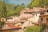 A picturesque village with a cluster of rustic houses topped with terracotta roofs. These homes are nestled among lush greenery and hills, creating a charming and serene landscape. The buildings are adorned with balconies and potted plants, and the sunlight casts a warm glow on the scene.