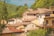 A picturesque village with a cluster of rustic houses topped with terracotta roofs. These homes are nestled among lush greenery and hills, creating a charming and serene landscape. The buildings are adorned with balconies and potted plants, and the sunlight casts a warm glow on the scene.