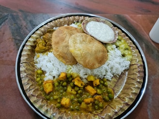 A plate with a generous serving of white rice, topped with two puris, surrounded by various curries including a pea and paneer curry, potato curry, and a side of raita in a small dish.