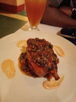 A plate of food featuring a richly sauced dish with herbs and spices on it. Sauces are artistically drizzled on the plate. A tall glass with a layered orange and yellow beverage sits in the background. The atmosphere suggests a dining setting.