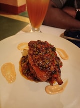 A plate of food featuring a richly sauced dish with herbs and spices on it. Sauces are artistically drizzled on the plate. A tall glass with a layered orange and yellow beverage sits in the background. The atmosphere suggests a dining setting.