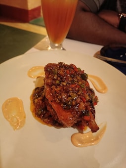 A plate of food featuring a richly sauced dish with herbs and spices on it. Sauces are artistically drizzled on the plate. A tall glass with a layered orange and yellow beverage sits in the background. The atmosphere suggests a dining setting.