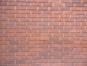A wall constructed with uniformly sized brownish-red bricks arranged in a horizontal pattern. The brick texture is visible, with slight variations in color and surface detailing on each brick.