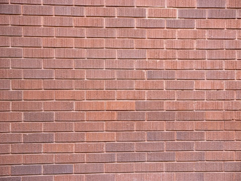 A wall constructed with uniformly sized brownish-red bricks arranged in a horizontal pattern. The brick texture is visible, with slight variations in color and surface detailing on each brick.