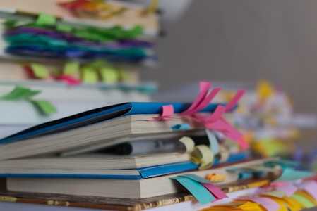 A close-up of several books stacked on top of each other, with colorful paper tabs sticking out from the pages. The tabs are in various colors like pink, blue, and yellow, adding a vibrant and organized appearance to the scene. The background is blurred, suggesting more books or similar objects.