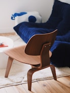 A modern, stylish wooden chair with a curved backrest is placed on a light, plush carpet. In the background, there is a deep blue couch that follows a contemporary design, situated near a wooden flooring. A cushion featuring blue and white accents rests on the couch. A white round table with a book is partially visible to the left.