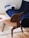 A modern, stylish wooden chair with a curved backrest is placed on a light, plush carpet. In the background, there is a deep blue couch that follows a contemporary design, situated near a wooden flooring. A cushion featuring blue and white accents rests on the couch. A white round table with a book is partially visible to the left.