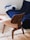 A modern, stylish wooden chair with a curved backrest is placed on a light, plush carpet. In the background, there is a deep blue couch that follows a contemporary design, situated near a wooden flooring. A cushion featuring blue and white accents rests on the couch. A white round table with a book is partially visible to the left.