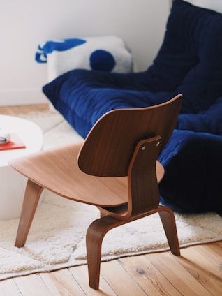A modern, stylish wooden chair with a curved backrest is placed on a light, plush carpet. In the background, there is a deep blue couch that follows a contemporary design, situated near a wooden flooring. A cushion featuring blue and white accents rests on the couch. A white round table with a book is partially visible to the left.