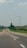 A straight road stretches into the distance with several cars driving along it. On the right side, a large wind turbine stands in a field, surrounded by patches of green grass and trees. The sky above is partly cloudy, creating a serene atmosphere.