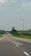 A straight road stretches into the distance with several cars driving along it. On the right side, a large wind turbine stands in a field, surrounded by patches of green grass and trees. The sky above is partly cloudy, creating a serene atmosphere.