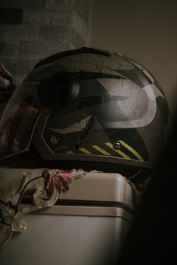 A motorcycle helmet with a camouflage pattern and a visor is resting on a white surface, partially covered by a floral-patterned fabric. The background features a textured wall with a muted, earthy tone.