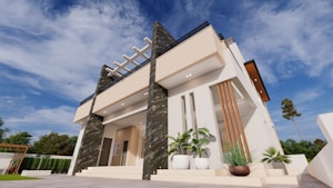 A modern two-story house with a flat roof, featuring a minimalist architectural style. The facade includes beige and dark textured panels, with large windows and a small terrace or balcony. Landscaping includes potted plants and surrounding greenery. The sky is bright and clear, suggesting a sunny day.
