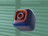 A 3D-rendered cube with a soft, smooth texture is floating against a blurred background. The cube is dark blue with an orange outline of a camera icon, resembling a popular social media logo.