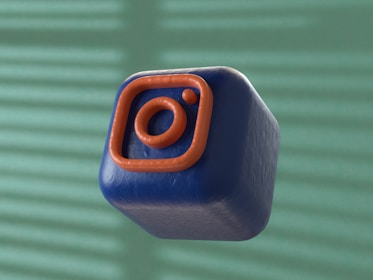 A 3D-rendered cube with a soft, smooth texture is floating against a blurred background. The cube is dark blue with an orange outline of a camera icon, resembling a popular social media logo.