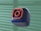A 3D-rendered cube with a soft, smooth texture is floating against a blurred background. The cube is dark blue with an orange outline of a camera icon, resembling a popular social media logo.