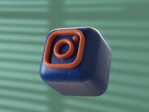 A 3D-rendered cube with a soft, smooth texture is floating against a blurred background. The cube is dark blue with an orange outline of a camera icon, resembling a popular social media logo.