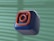 A 3D-rendered cube with a soft, smooth texture is floating against a blurred background. The cube is dark blue with an orange outline of a camera icon, resembling a popular social media logo.
