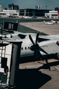 A turboprop aircraft is parked at an airport gate, with its propeller prominently visible in the foreground. The setting includes a tarmac with a boarding bridge labeled B1 and several service vehicles scattered across the area. In the background, a modern terminal building and distant city skyline are visible under a clear blue sky.