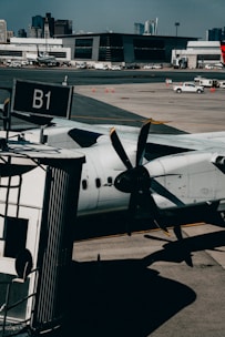A turboprop aircraft is parked at an airport gate, with its propeller prominently visible in the foreground. The setting includes a tarmac with a boarding bridge labeled B1 and several service vehicles scattered across the area. In the background, a modern terminal building and distant city skyline are visible under a clear blue sky.