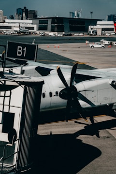 A turboprop aircraft is parked at an airport gate, with its propeller prominently visible in the foreground. The setting includes a tarmac with a boarding bridge labeled B1 and several service vehicles scattered across the area. In the background, a modern terminal building and distant city skyline are visible under a clear blue sky.
