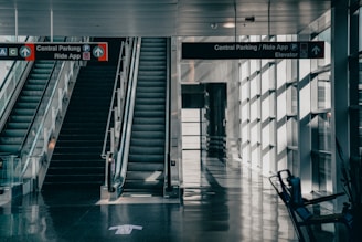 A modern airport corridor featuring two parallel escalators with signage above indicating directions to central parking and ride apps. The area is brightly lit by large windows on the right, casting reflections and shadows on the polished dark floor.