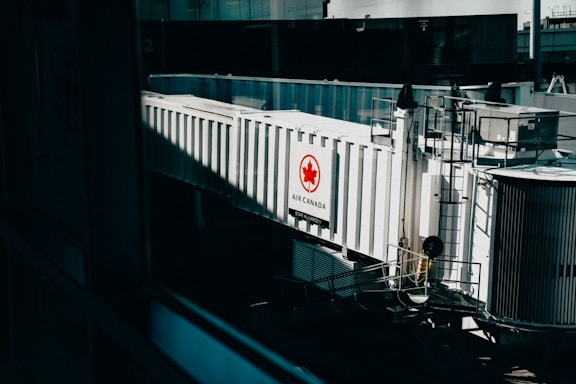 A jet bridge connected to an Air Canada terminal, featuring a prominent Air Canada logo on the side. The structure is white with glass panels and railings visible, set in an airport environment with a hint of industrial design.
