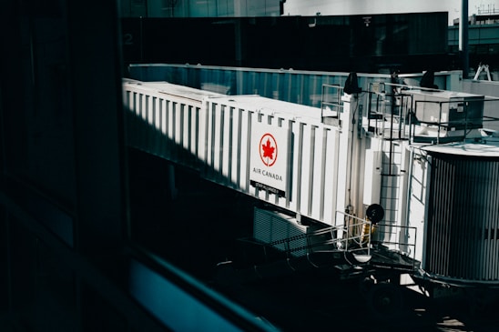 A jet bridge connected to an Air Canada terminal, featuring a prominent Air Canada logo on the side. The structure is white with glass panels and railings visible, set in an airport environment with a hint of industrial design.