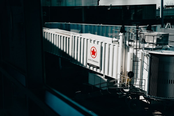 A jet bridge connected to an Air Canada terminal, featuring a prominent Air Canada logo on the side. The structure is white with glass panels and railings visible, set in an airport environment with a hint of industrial design.