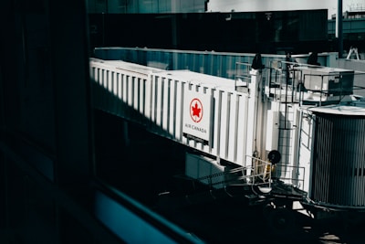A jet bridge connected to an Air Canada terminal, featuring a prominent Air Canada logo on the side. The structure is white with glass panels and railings visible, set in an airport environment with a hint of industrial design.