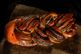 A braided loaf of bread with a glossy, golden-brown crust placed on a piece of burlap. The bread appears to be enriched with dark, possibly poppy seed filling within the folds.