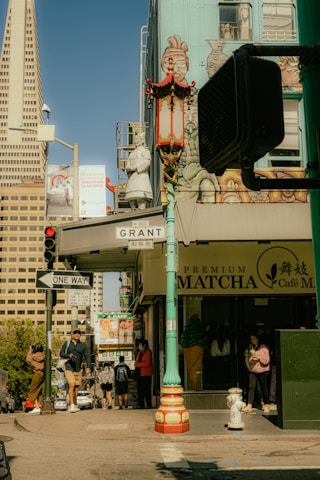 A city street corner with signs for Grant Avenue and a one-way street is bustling with pedestrians. The street features a mural with an Asian motif, a red and gold lamppost with decorative elements, and an ice cream shop called Premium Matcha Café. The Transamerica Pyramid is visible in the background, indicating a location in San Francisco's Chinatown. People are walking in various directions and engaging in activities like checking their phones.