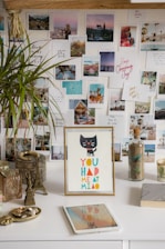 A wall is decorated with a collage of various photographs, featuring landscapes, inspirational quotes, and people in different settings. A tabletop holds a framed illustration of a black cat with the text 'YOU HAD ME AT MIAO' in colorful letters. Nearby are decorative items such as a small Eiffel Tower model, candles, plants, and glass bottles filled with natural elements. An electronic tablet lies flat on the surface.