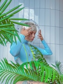 A person with short blond hair wearing a futuristic, light blue outfit and helmet. The helmet is designed to resemble an astronaut's headgear and is labeled with the word 'ALIEN'. The person is adjusting the helmet while surrounded by green tropical plants. The background consists of white tiled walls, giving a clean, modern look.