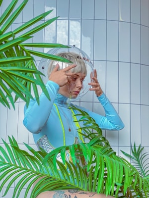 A person with short blond hair wearing a futuristic, light blue outfit and helmet. The helmet is designed to resemble an astronaut's headgear and is labeled with the word 'ALIEN'. The person is adjusting the helmet while surrounded by green tropical plants. The background consists of white tiled walls, giving a clean, modern look.