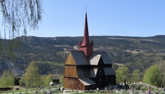 A scenic landscape featuring a medieval wooden stave church with a red pointed steeple and dark timber walls, surrounded by a lush green cemetery. The backdrop includes rolling forested hills and a clear blue sky, with some distant mountains visible.