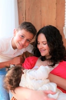 A woman with curly dark hair wearing a pink top is sitting and smiling at a baby wearing a cream outfit in her arms. A young boy with short brown hair and a white shirt leans in from the side, smiling at the camera. They are seated against a wooden wall with a soft white curtain visible.