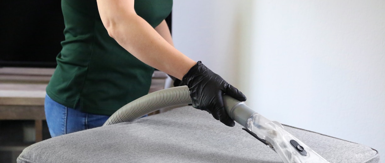 a woman in a green shirt and black gloves vacuuming a gray ottoman