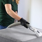 a woman in a green shirt and black gloves vacuuming a gray ottoman